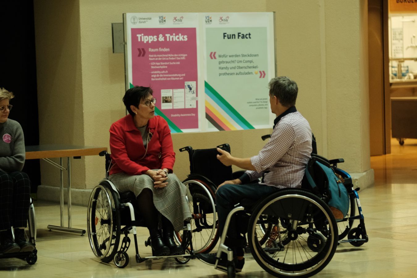 Rollstuhlsimulation: Zwei Personen im Rollstuhl, darunter die Prorektorin Lehre und Studium, Gabriele Siegert, sprechen miteinander vor Plakaten mit Tipps und Informationen zur Barrierefreiheit im Lichthof der UZH.